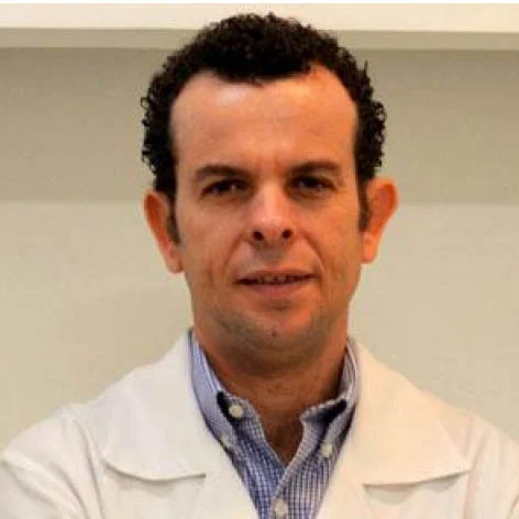 Médico Rafael Gualberto, visto de frente, com jaleco branco, e camisa social de listras azuis por de baixo.